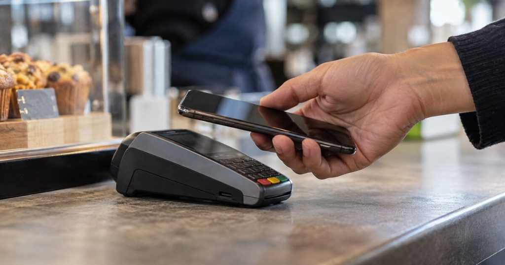 A hand holding a smartphone above a contactless payment terminal at a cafe.